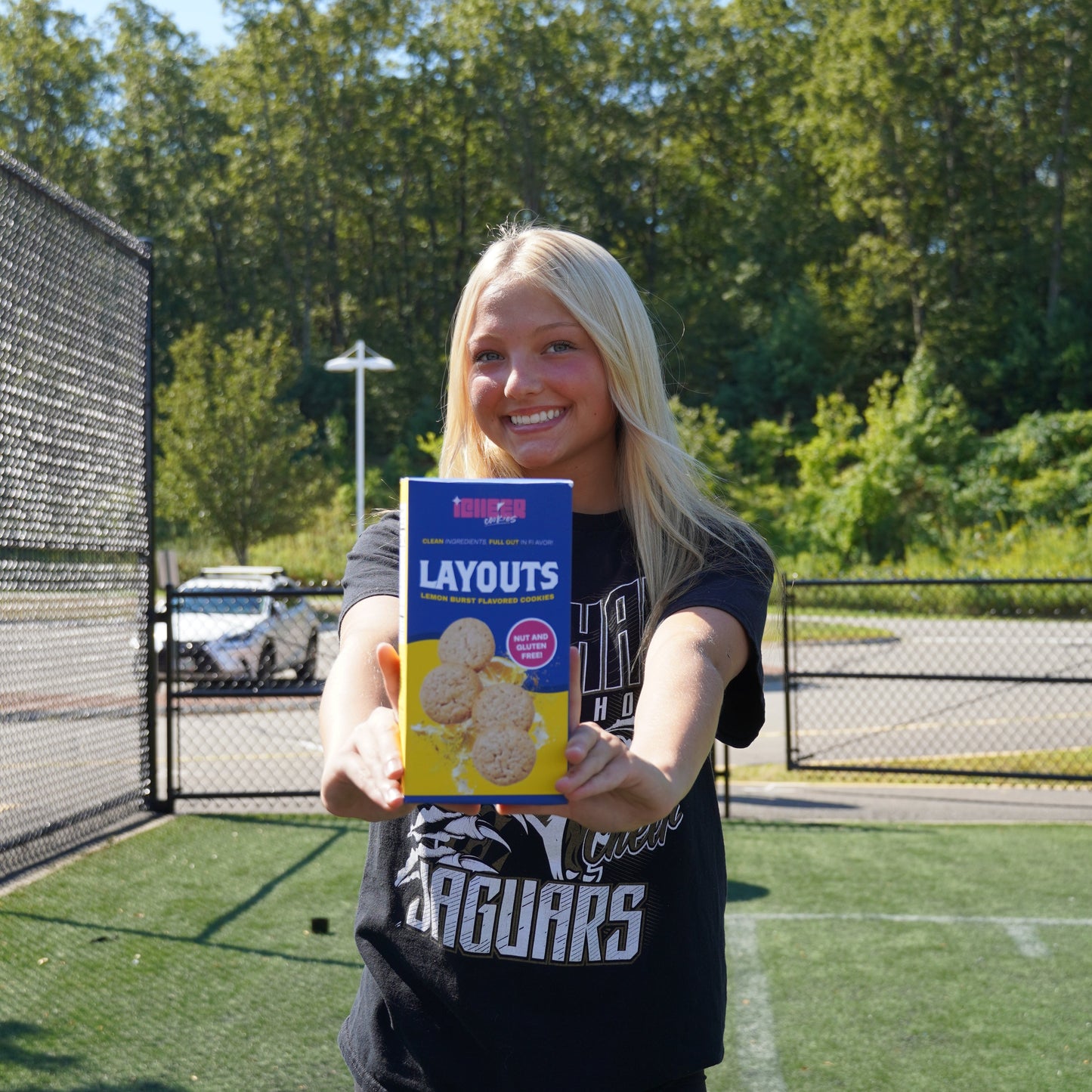 Person holding a box of Lemon  iCheer cookies on a sports field with trees and clear sky in the background