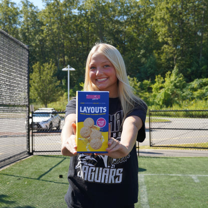 Person holding a box of Lemon  iCheer cookies on a sports field with trees and clear sky in the background